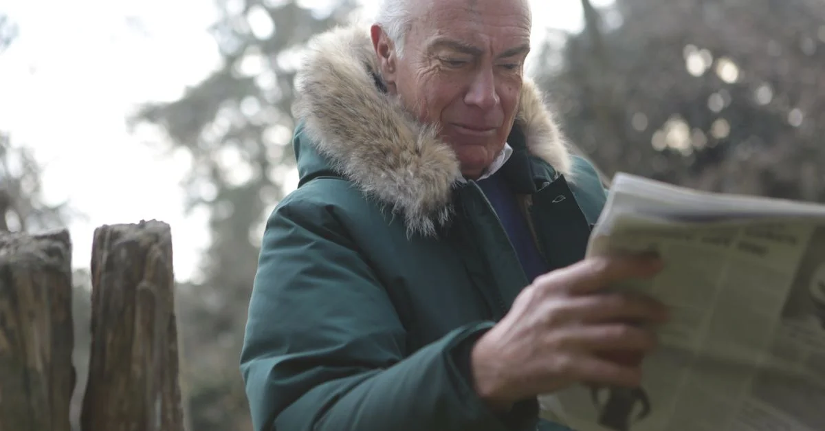 Un homme âgé en manteau d'hiver lit un journal dans un parc, capturant un moment de détente.