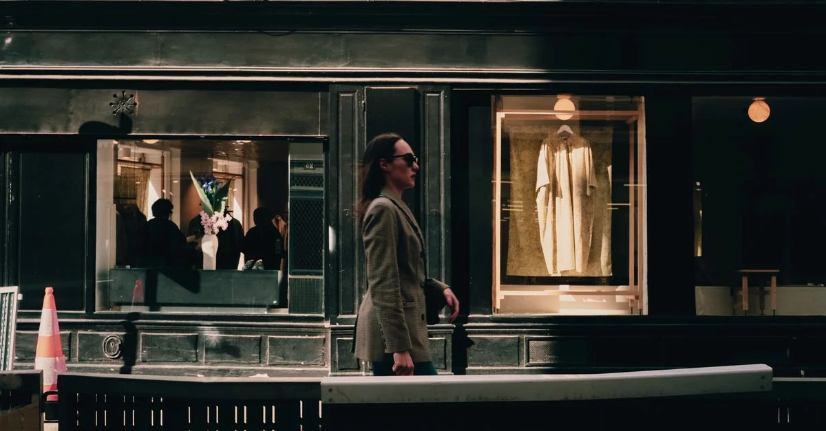 Une femme à la mode en blazer passe devant une vitrine urbaine stylée par une journée ensoleillée.